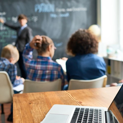 Laptap auf Tisch mit Teilnehmenden, Tafel und Dozent